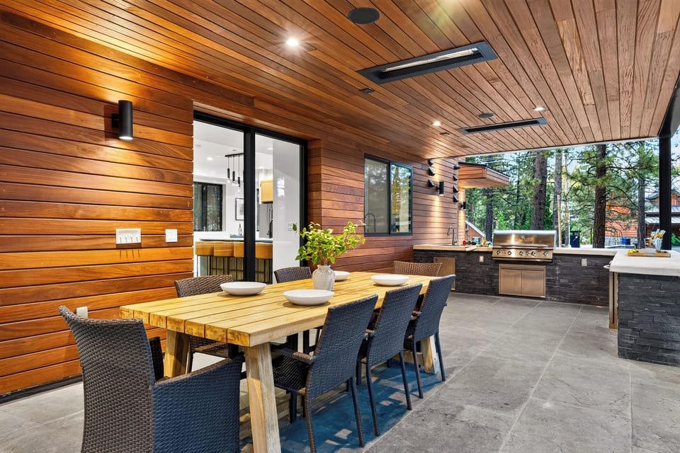 Covered outdoor dining area with a built-in grill, warm wood finishes, and forest views.