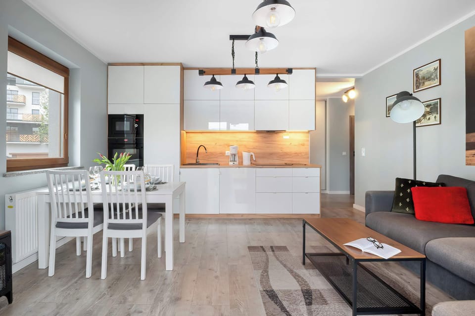 A wide view of the living room highlights the seamless flow between the dining table, the kitchen, and the lounge area. Decorative lighting fixtures hang from the ceiling, adding a touch of industrial style to the bright interior.