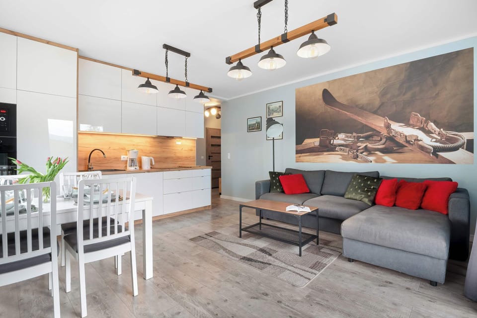 An open-concept living area combines a sleek white kitchen with a comfortable gray sectional sofa adorned with red accent pillows. A wooden dining table sits in the foreground, creating a functional and modern living space.