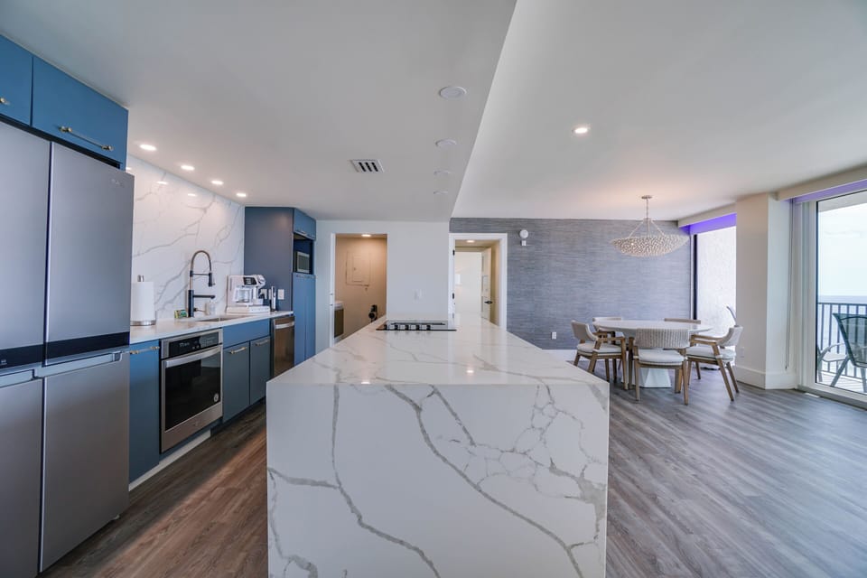 Your modern open-plan living space flows seamlessly from a sleek blue kitchen with marble counters to a stylish dining area, with ocean views just beyond the balcony doors.