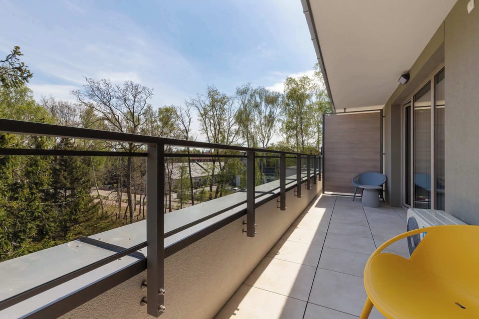 A spacious balcony offers a view of lush green trees behind a dark metal railing. The outdoor flooring is light gray tile, and a bright yellow modern chair sits in the corner for relaxation.