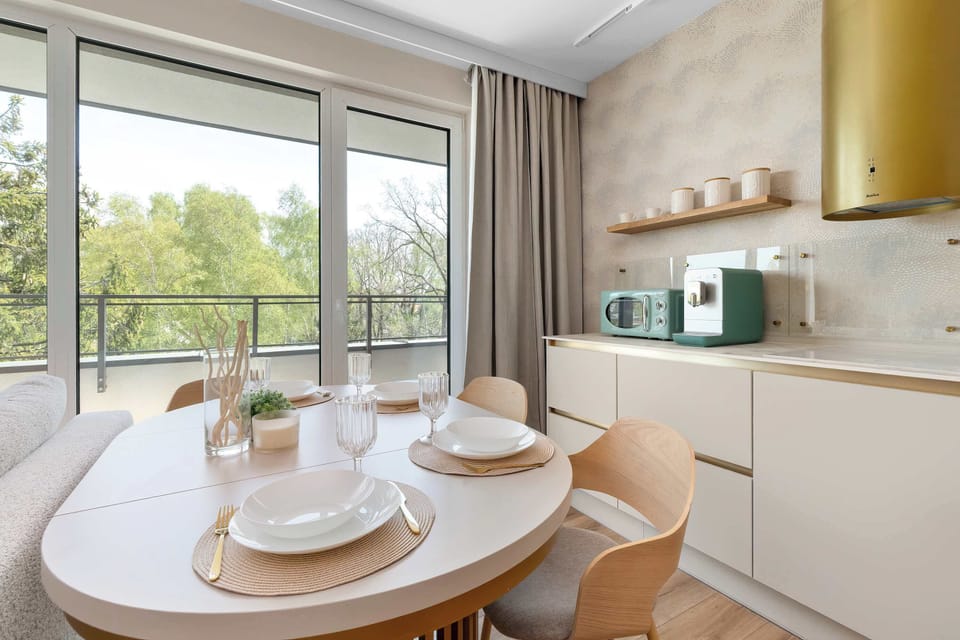 The dining area is captured from an angle showing a round table set with elegant white plates and gold-rimmed glasses. Large windows in the background provide a seamless transition to the outdoor greenery.