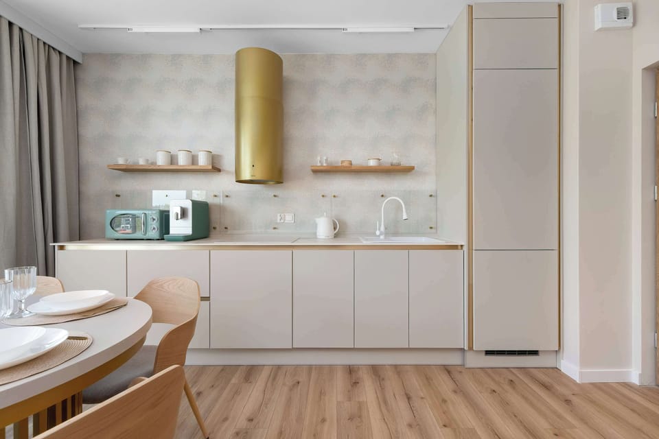 A sleek kitchenette wall showcases light-colored cabinetry paired with a distinctive gold cylindrical extractor hood. The countertop is neatly organized with a coffee machine and small kitchen accessories.