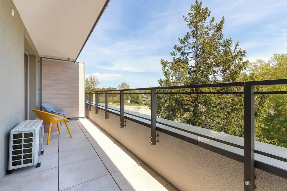 The length of the balcony shows the glass and metal railing stretching toward a view of the sky and trees. An air conditioning unit is discreetly placed on the tiled floor near the corner.