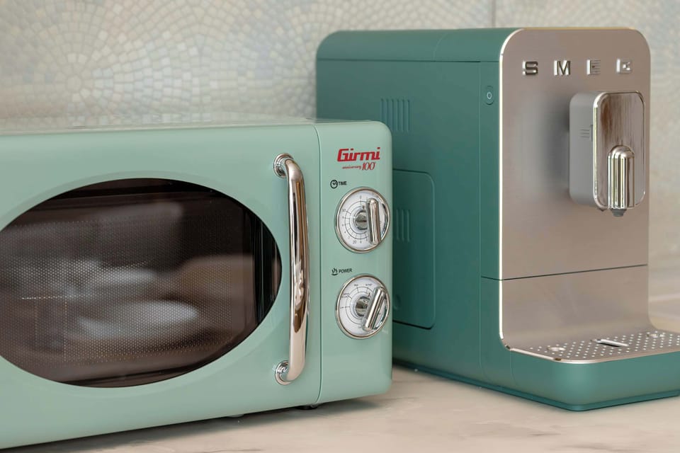 A close-up of kitchen appliances features a retro-style mint green toaster and a modern dark teal espresso machine. These high-end appliances sit on a light-colored countertop against a subtly patterned backsplash.