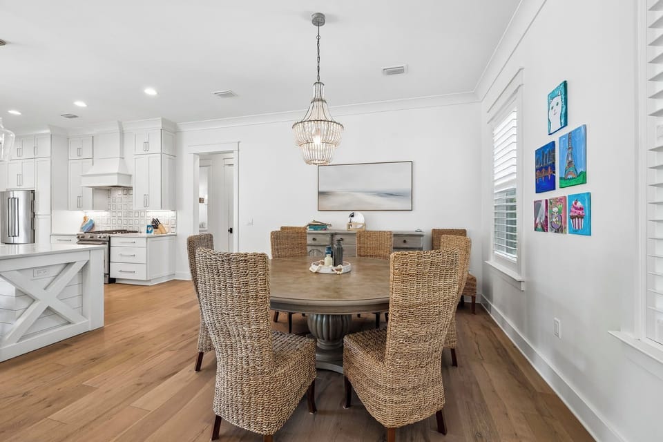 Gather around this inviting dining space where coastal elegance meets everyday comfort. Your morning coffee or sunset dinner feels special beneath the statement chandelier, with woven chairs and warm wood tones creating a welcoming atmosphere.