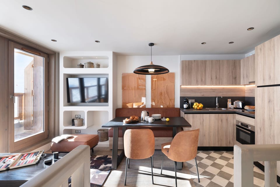 Dining area with wall-mounted TV, black table, and warm wood chairs — bright natural light from the windows
