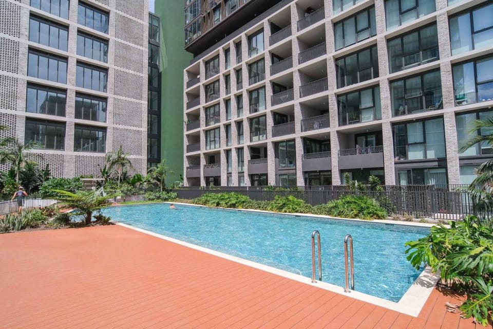 The building`s shared outdoor swimming pool from the deck end, showing the wide terracotta coloured surround and the stainless steel entry ladder. Tropical garden beds line all sides.