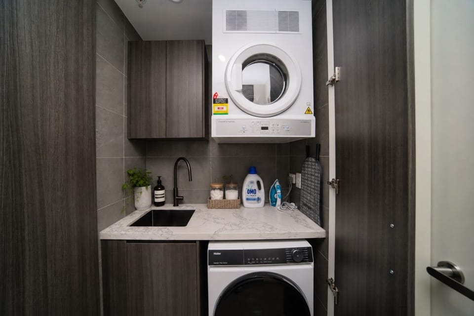 The laundry with a Haier front load washing machine at floor level and a Fisher and Paykel dryer mounted above. A marble effect benchtop and small undermount sink sit alongside, with an iron and ironing board stored here.