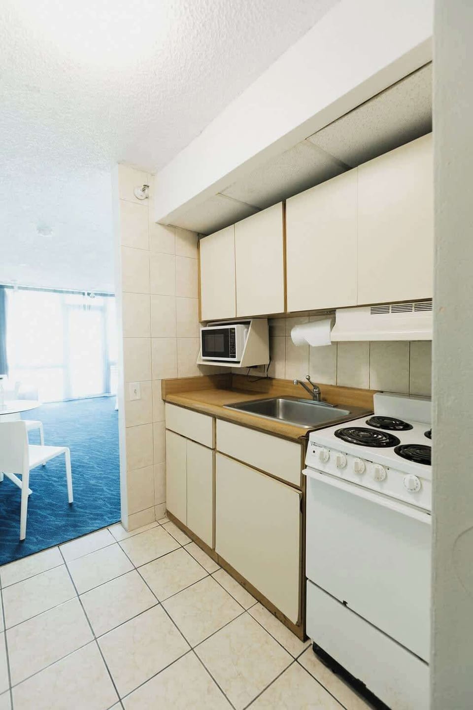 Compact vintage kitchenette with electric stove, sink, overhead cabinets, and microwave, opening into the bright studio for an efficient home‑style setup.