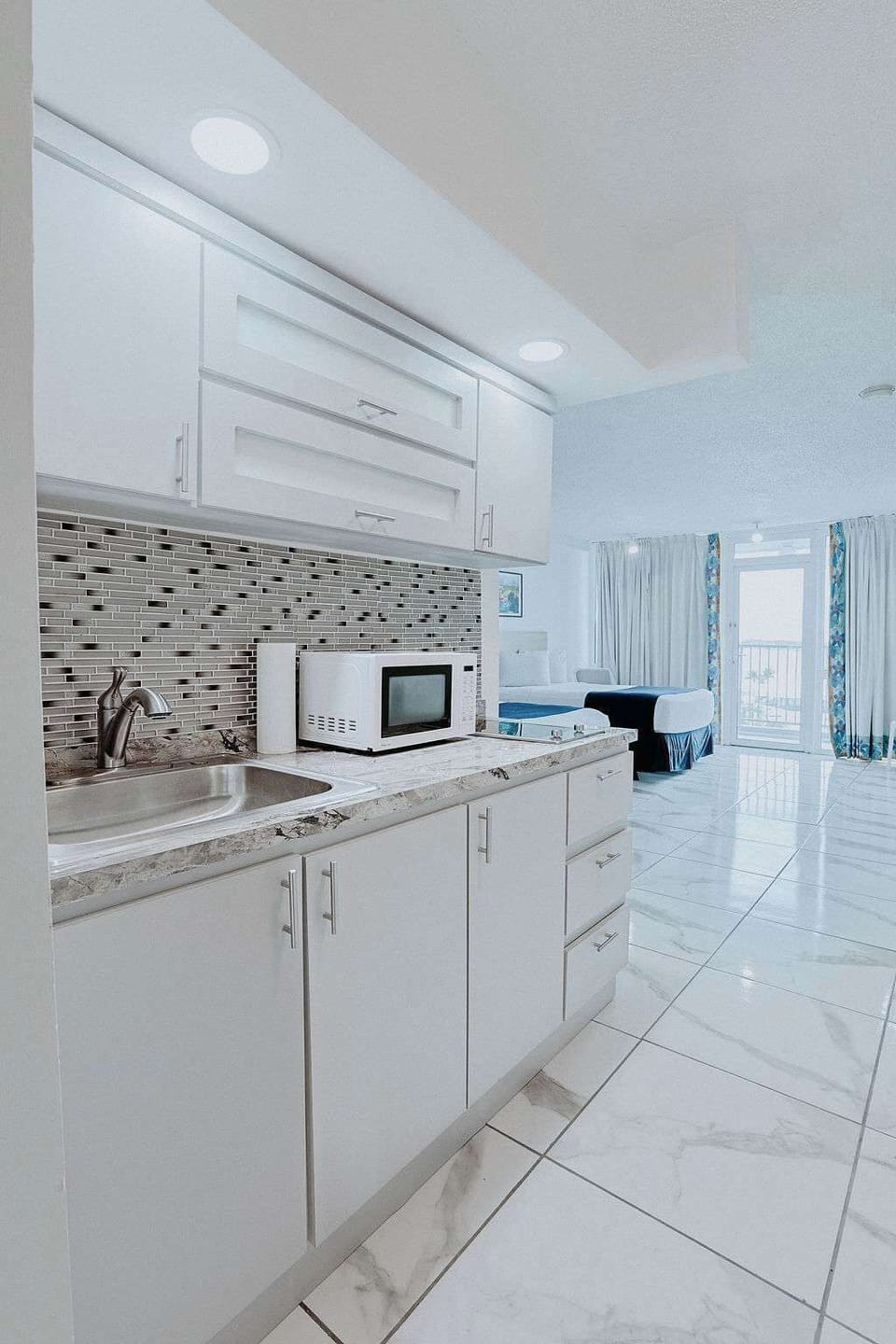 Sleek white kitchenette with marble‑style counters, glass tile backsplash, stainless sink, and bright recessed lighting, opening directly into your double‑bed studio with balcony.
