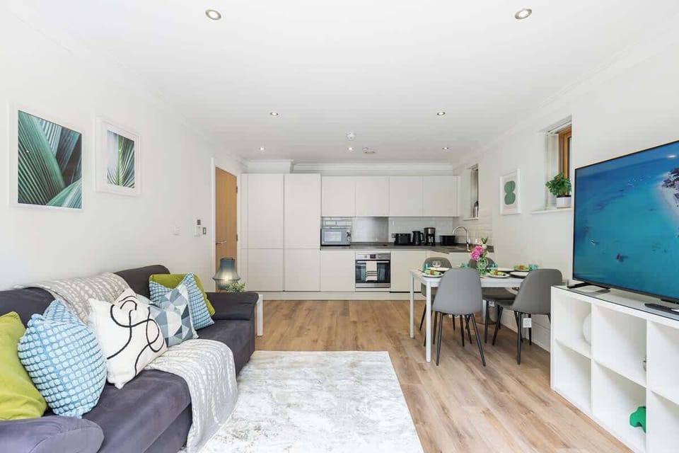 Relax and dine in this spacious open-plan living area, featuring a dark sofa styled with colorful throw pillows, a set dining table with grey chairs, a modern white kitchen, and a large flat-screen TV set above warm wooden flooring.