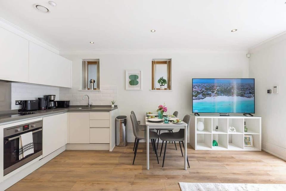 Gather and dine in this bright, open-plan living space, featuring a modern handleless kitchen with integrated appliances, a dining table set for four, and a large flat-screen TV resting on a white cube storage unit over warm wooden flooring.