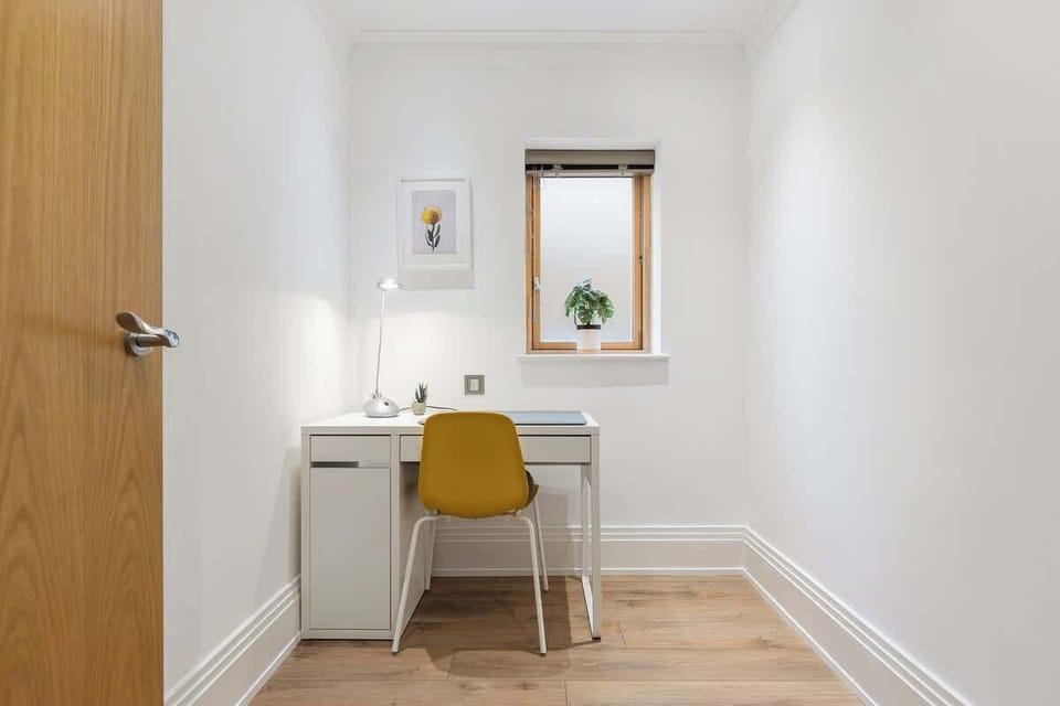 Focus or work in this quiet home office nook, featuring a white writing desk, a mustard yellow chair, a desk lamp, light wooden flooring, and a small window allowing natural light.
