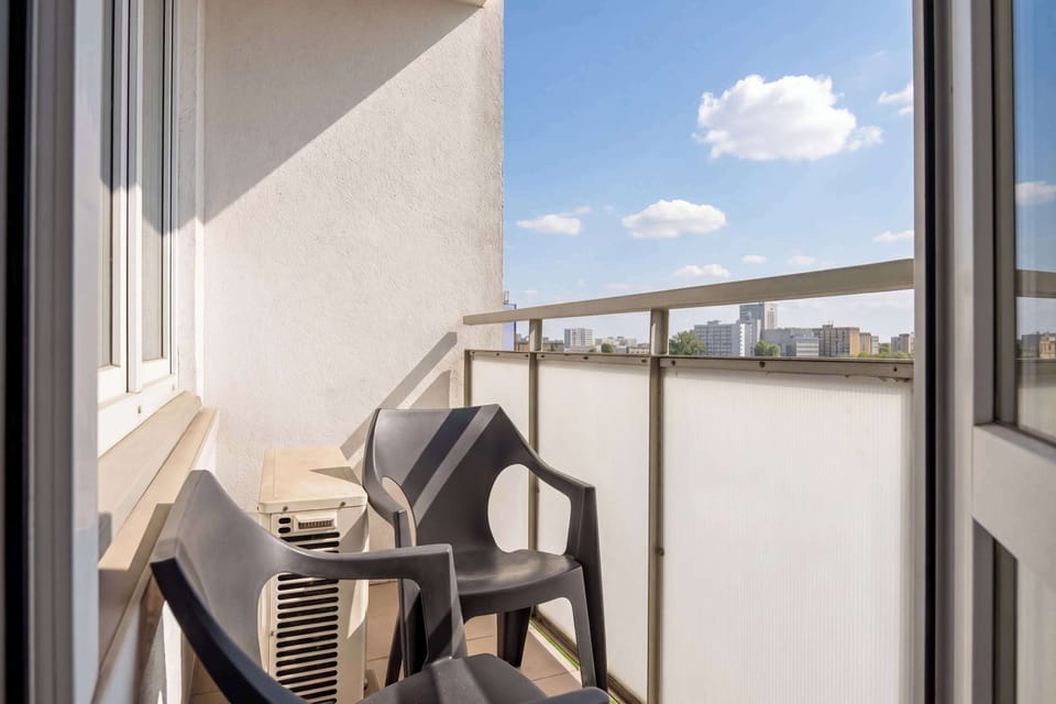 A sunny balcony is equipped with dark outdoor chairs, offering a private spot for relaxation. A metal railing secures the space, which overlooks the surrounding area under a clear blue sky.