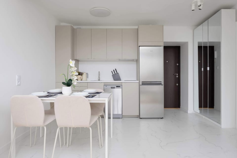 A dining area features a white table set for a meal, surrounded by four upholstered chairs, positioned right next to a modern kitchen zone. A large mirrored wardrobe stands in the hallway nearby, reflecting the light space.