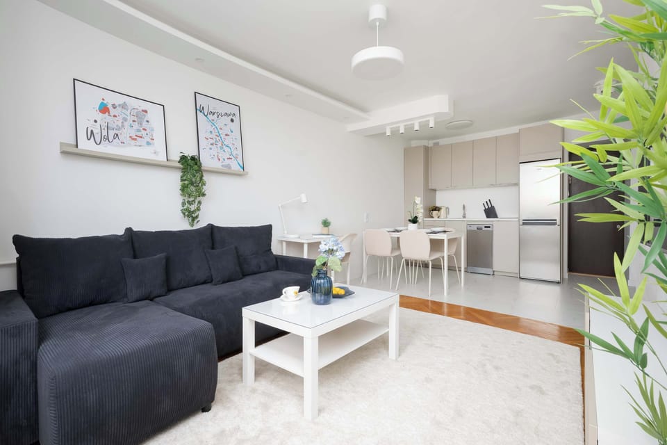 An open-concept living space combines a dark corner sofa, a low white coffee table, and a dining table set further back. Potted green plants add a touch of freshness to the bright interior.