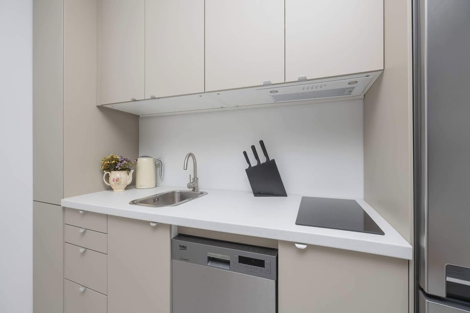 This compact kitchen corner includes sleek white cabinetry, a built-in cooktop, a sink with a modern faucet, and a dishwasher. A block of kitchen knives sits neatly on the countertop next to a stainless steel refrigerator.