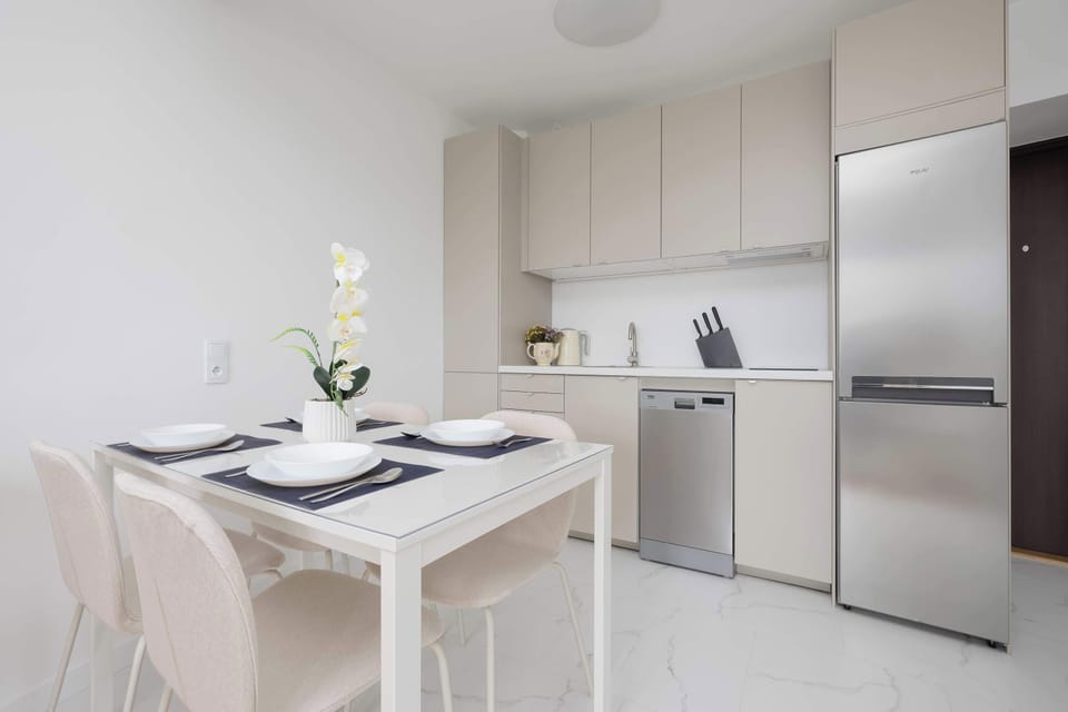 The kitchen and dining space seamlessly connect, showcasing a white table set for four and sleek kitchen cabinets in the background. A stainless steel refrigerator completes the functional, modern design.