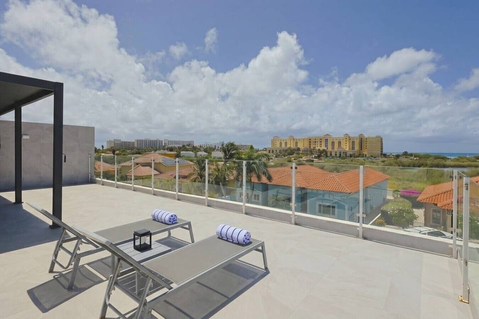 Tiled rooftop terrace equipped with two sun loungers and glass railings, offering an elevated view of a nearby resort.