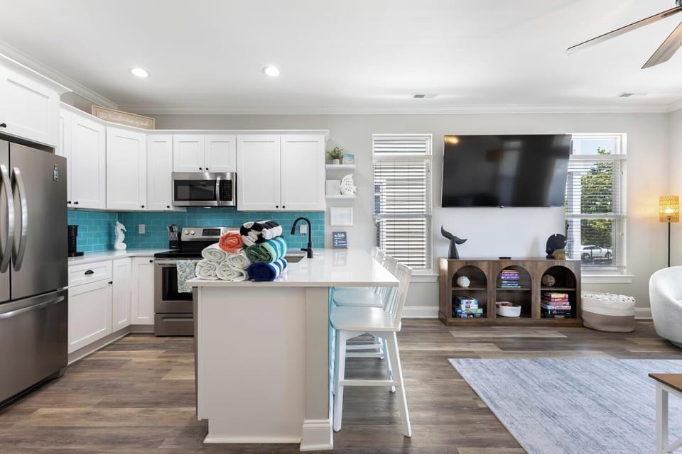Bright open-concept space with a fully-equipped kitchen featuring coastal blue backsplash, island seating, stainless appliances, and living area with wall-mounted TV—your perfect home base.