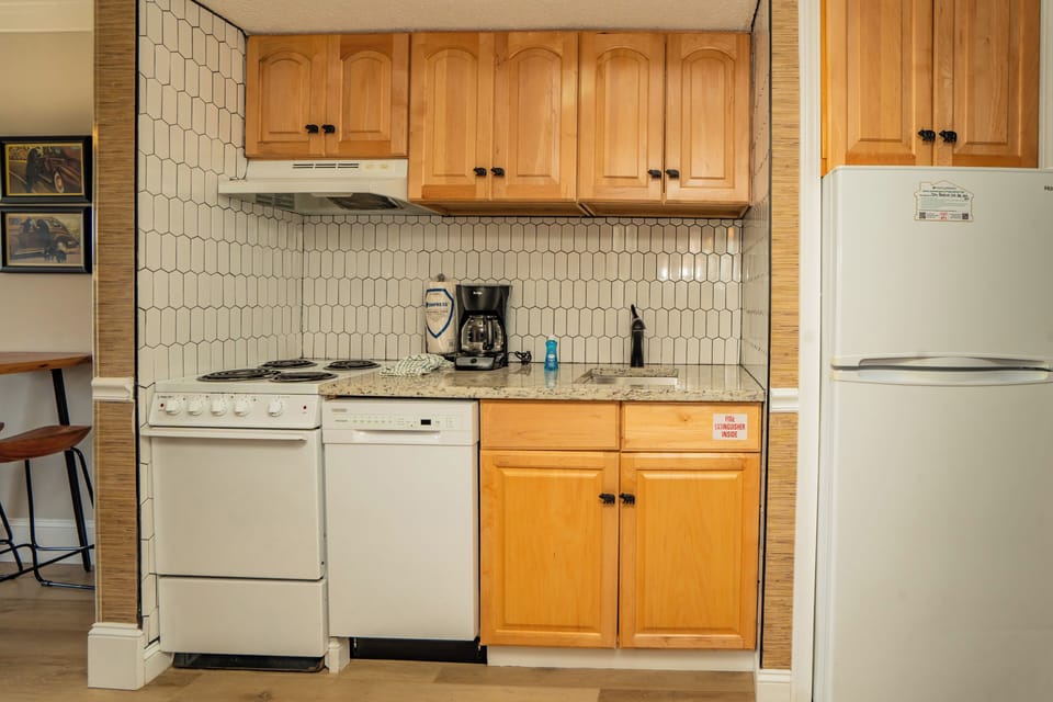 Your kitchenette is equipped with everything you need to prepare meals—a stove, dishwasher, refrigerator, and coffee maker ready for your morning brew. Warm wood cabinetry and charming tile backsplash add a cozy, welcoming touch.