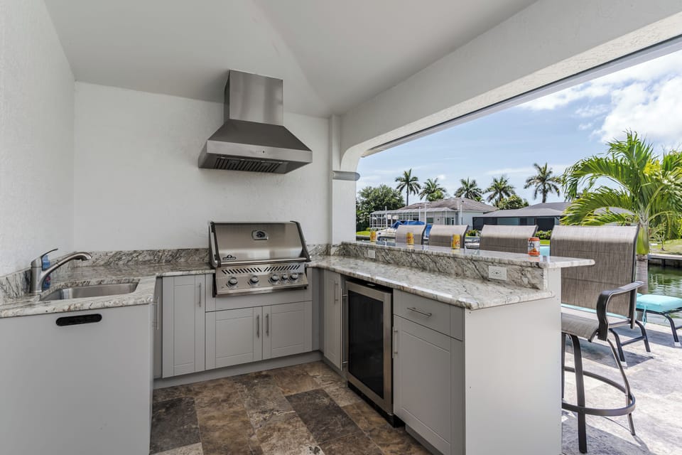 Your private outdoor kitchen features a built-in grill, bar seating, and refreshing canal views framed by swaying palms—perfect for sunset meals by the pool.