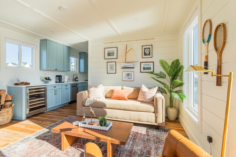 The living area looking toward the galley kitchen, with the maritime gallery wall, queen sleeper sofa, vintage tennis rackets, brass accents, and the wine fridge visible from across the open-plan space.