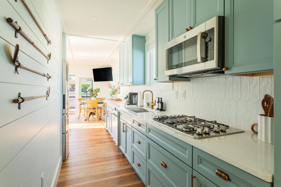 The galley kitchen looking toward the dining area, with blue-gray cabinetry, brass hardware, a gas cooktop, quartz counters, and a white geometric tile backsplash running the full length of the space.
