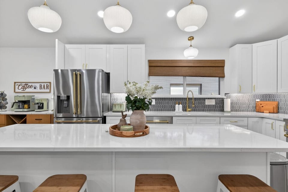 Spacious gourmet kitchen with a large quartz island, seating for four, and a stainless French-door fridge with brass accents. Designer pendant lighting, custom picket backsplash, white shaker cabinets, and a dedicated coffee bar nearby