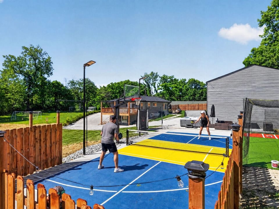 Sunny afternoon pickleball match on the private blue and yellow court. Just beyond the net you`ll spot the hot tub, basketball hoop, soccer goal, and batting cage—your own backyard sports complex ready for the whole crew to enjoy.