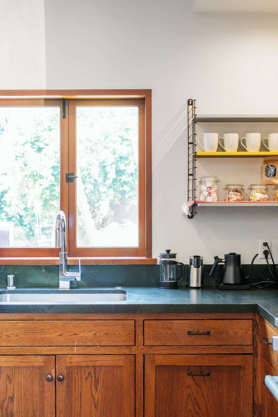 Kitchen — Close-Up of Undermount Stainless Sink, Chrome Gooseneck Faucet, Dark Green Soapstone Counter, Garden-View Casement Window, Wire Shelving with White Mugs & Coffee and Tea Station