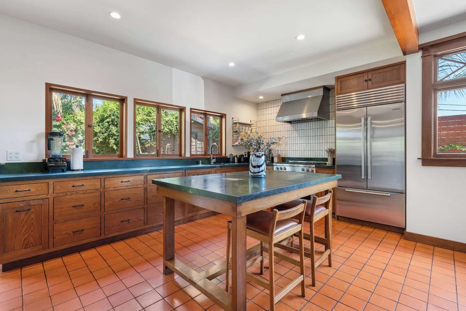 Kitchen — Wide View Showing Oak Cabinets, Dark Green Soapstone Counters, Center Island with 2 Leather Bar Stools, Terracotta Tile Floor, Stainless Range with Hood, Built-In Refrigerator & White Grid Tile Backsplash