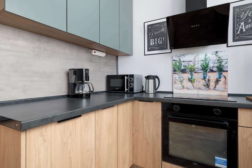 The sleek kitchen corner showcases minimalist light-blue cabinetry paired with warm wood-textured lower units. Modern appliances like a black microwave and an electric kettle are neatly arranged on the dark countertop.