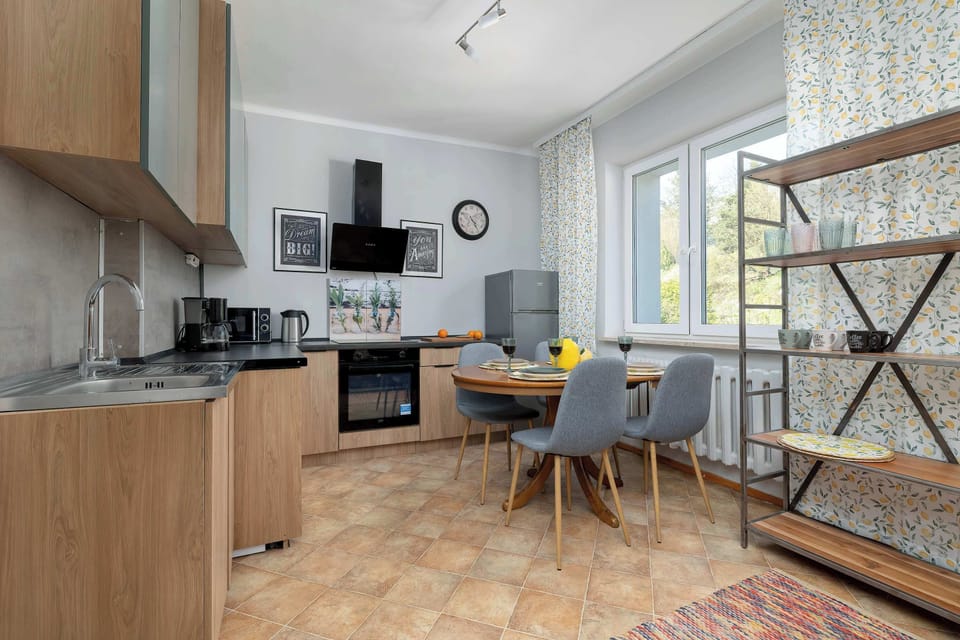 A modern kitchen and dining space features a minimalist wooden table surrounded by stylish grey upholstered chairs. Large windows look out onto greenery, creating a pleasant environment for meals.