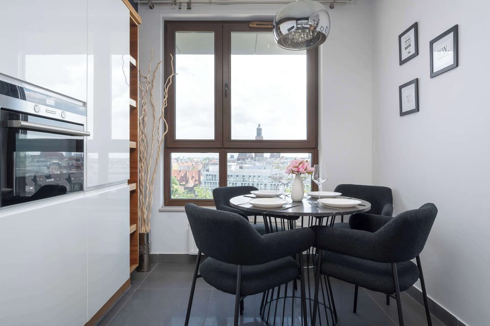 A central view of the dining area shows four black upholstered chairs arranged around a dark table. The space is illuminated by natural light from the large window, making it an inviting spot for meals.