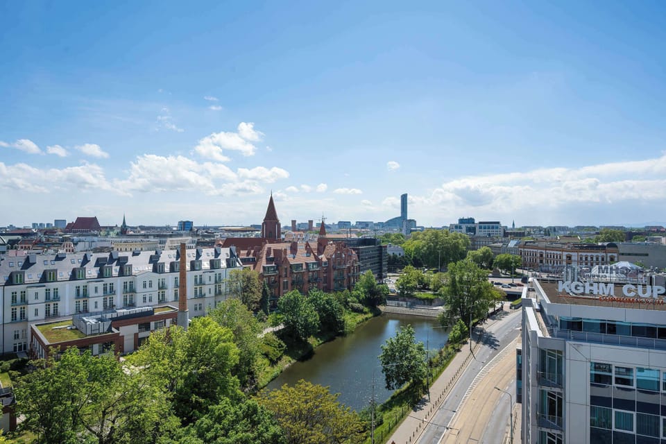 A view of the river and park showcases a historic red-brick building reflected in the calm water. Mature trees line the paths, making it an ideal neighborhood for nature lovers.