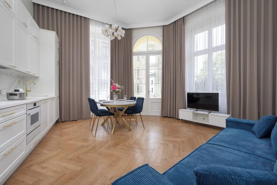 A spacious living room with parquet flooring, tall windows, and a blue sectional sofa.