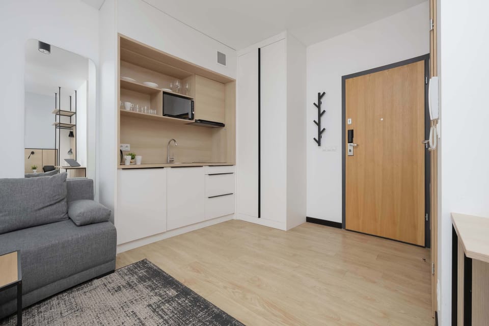A spacious view of the apartment showing the kitchenette, entrance door, and part of the living area with a sofa. Light wood flooring and white walls keep the room bright and modern.