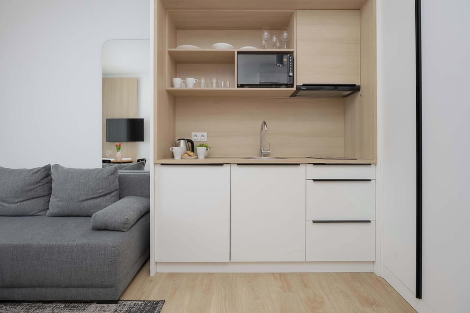 A sleek kitchen area with white cabinetry, a microwave, and wooden shelving above the counter. The space appears functional and neatly organized.