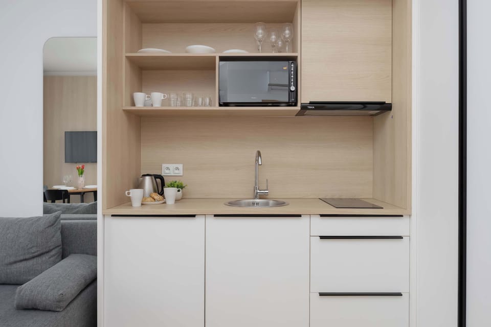 A close-up view of a modern kitchenette with white lower cabinets, wooden shelves, a microwave, and a compact sink area. Minimal appliances and tidy surfaces emphasize the simple design.