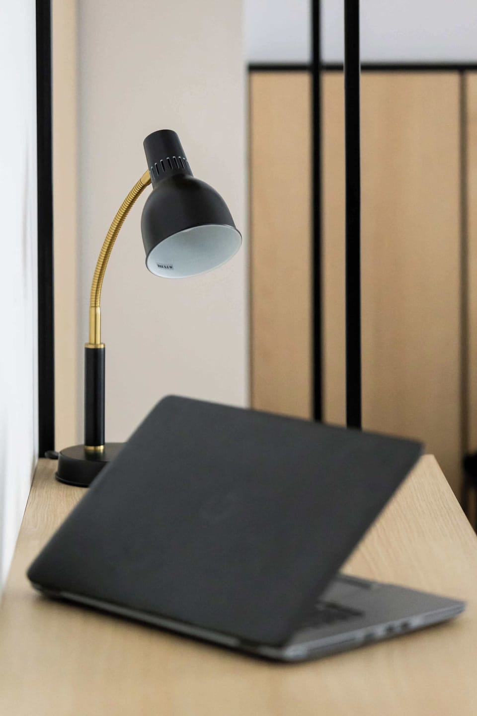 A close-up shot of a black desk lamp illuminating a dark notebook placed on a wooden desk. The blurred background highlights the workspace details.