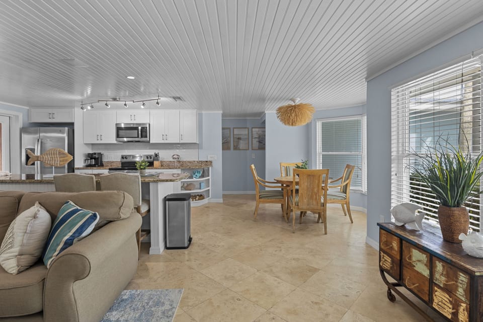 Step into a bright, coastal-inspired open living space where you can relax on comfortable seating, enjoy meals at the dining table, and prepare snacks in the well-equipped kitchen—all while soaking in natural light and beachy charm.