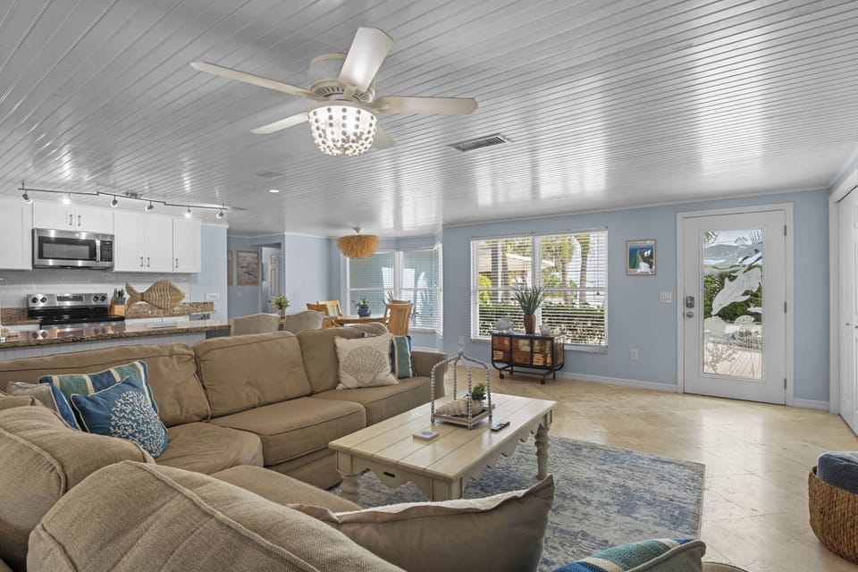 Relax in your spacious living room, where coastal charm meets comfort. Soft light filters through large windows while a gentle ceiling fan circulates the breeze above plush seating perfect for gathering with loved ones.