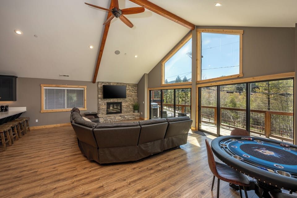 Your living space features soaring vaulted ceilings with warm wood beams, a cozy stone fireplace, and floor-to-ceiling windows framing peaceful forest views—ideal for relaxing or gathering with loved ones.