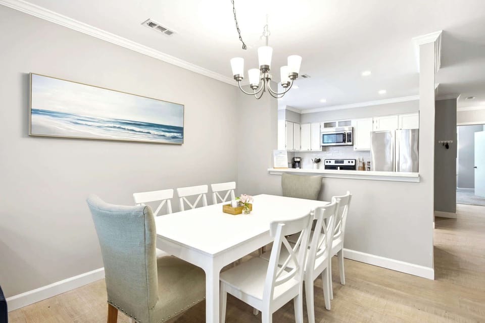 Crisp white furniture and coastal art create a serene setting for morning coffee or shared meals. This airy dining space flows seamlessly into a modern kitchen, glowing under the gentle radiance of a classic chandelier.