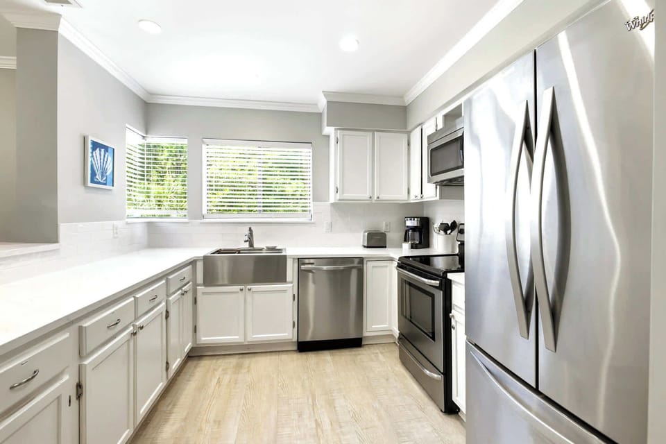 Polished stainless steel and crisp white cabinetry create a luminous, modern space for morning coffee. Natural light filters through the blinds, highlighting the sleek apron sink and airy layout designed for effortless coastal cooking.