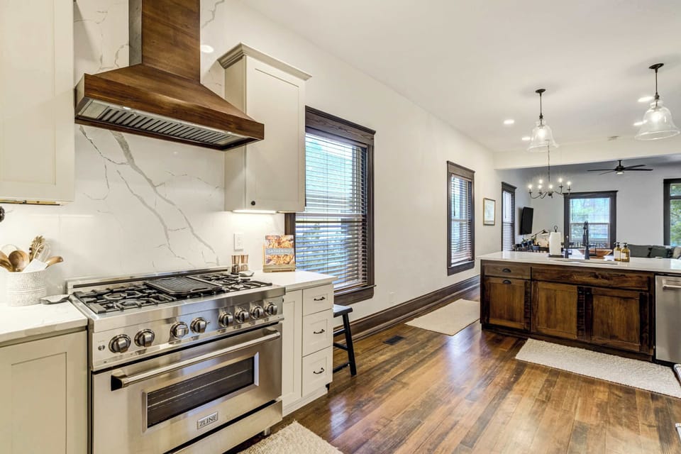 A professional-grade gas range sits beneath a custom wood-paneled range hood, with marble-look countertops and white shaker cabinetry framing a kitchen built for serious cooking and effortless entertaining.