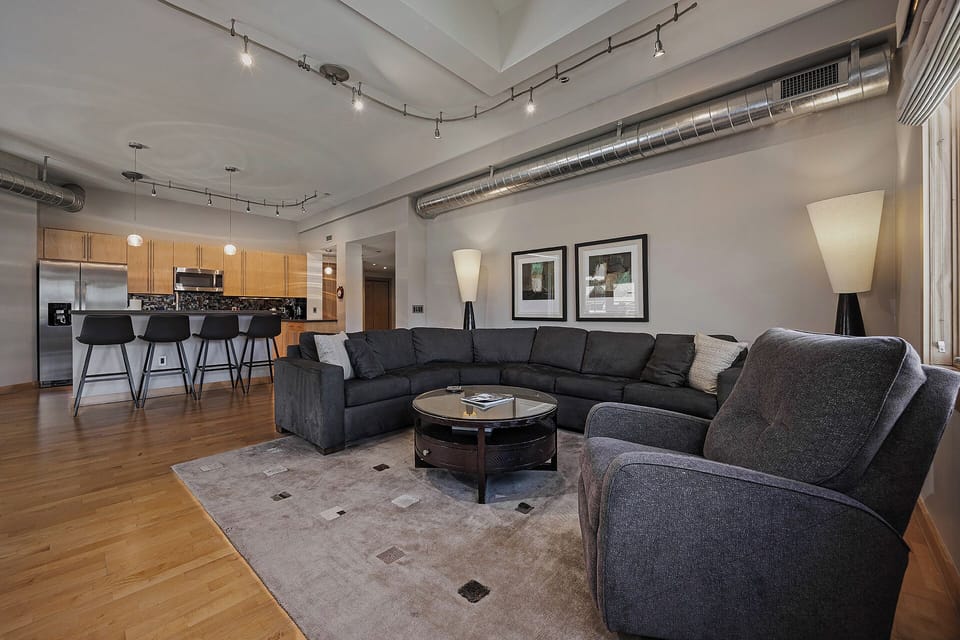 This modern, open-concept space features exposed ductwork and abundant natural light. A spacious L-shaped sectional anchors the living area, while the fully equipped kitchen with bar seating invites you to prepare meals and gather with loved ones.