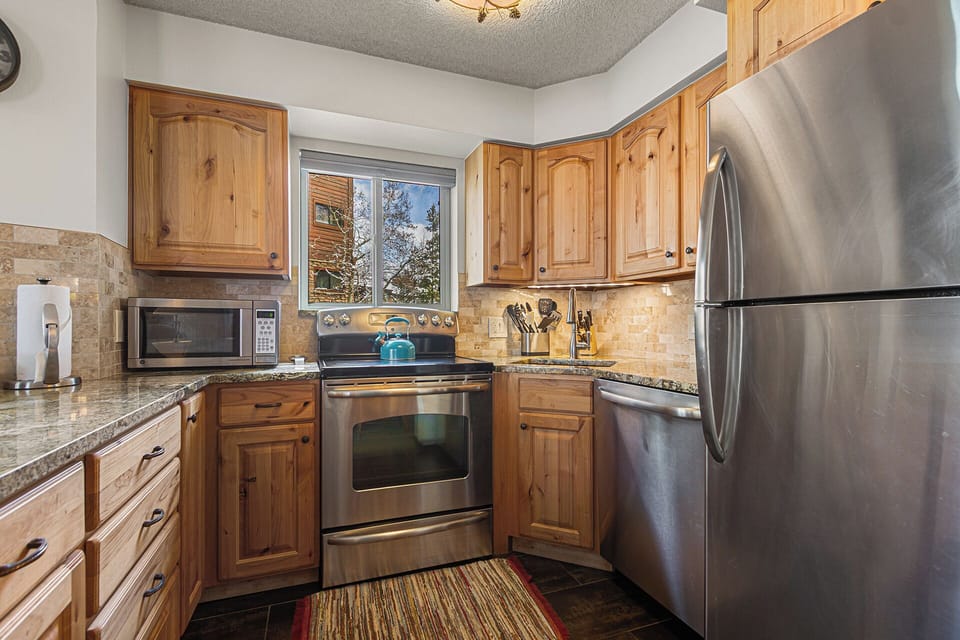 Your fully equipped kitchen awaits with warm wood cabinetry, stainless steel appliances, granite countertops, and mountain views through the window—perfect for preparing home-cooked meals during your stay.